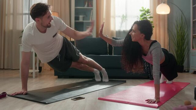 Multiethnic family happy couple Hispanic girl woman female exercising with Caucasian male man guy planking push-up plank practicing sport balance giving high five success teamwork inside living room