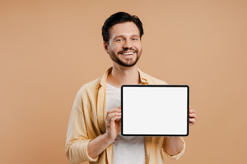 Smiling, bearded man showing digital tablet with empty screen on beige background looking at camera