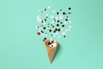 Shiny golden confetti flying out of ice cream cone on turquoise background, top view