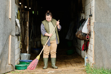 Person standing at barn entrance holding broom and sweeping wooden floor. Wearing warm layers and...