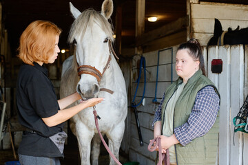 Two women engaging in conversation about horse care in stable setting with white horse standing...