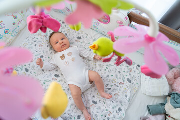 A baby lies on a vibrant play mat, exploring soft toys and colorful hanging flowers in a cozy, well-lit nursery