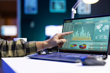 An individual pointing at a screen that shows numbers and financial charts related to stock trading. At a desk, digital laptop is displaying graphs and values for forex trends.