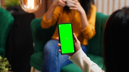 Woman checking greenscreen display on smartphone layout, passing time in lounge area at hotel. Asian tourist sitting on sofa with phone running isolated chromakey template on screen.