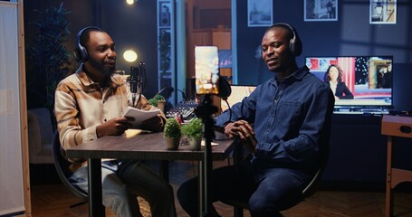 Show host talking with guest in studio, listening to opinion on current events, filming video for social media using smartphone on tripod. Influencer recording podcast with mobile phone