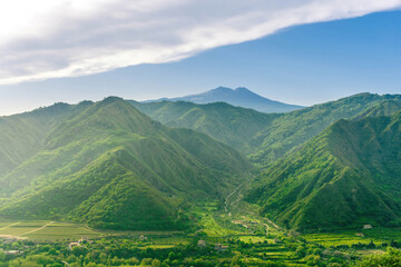 amazing view at a green landscape between two mountains with a high peak with snow and blue cloudy sky on background