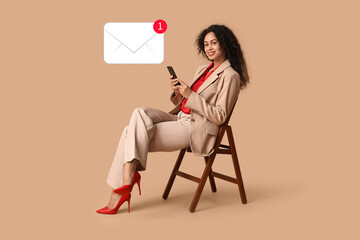 Beautiful young happy African-American woman in stylish suit with mobile phone sitting on chair against beige background