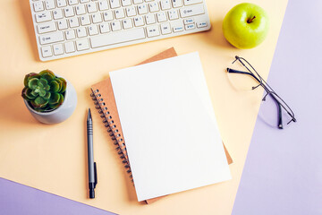 Desktop with blank paper, notepad, computer keyboard, glasses and green apple on beige and purple background. Top view, flat lay, mockup