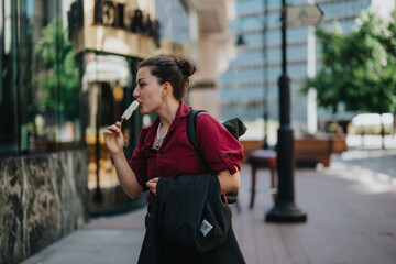 Fototapeta premium Young businesswoman savoring ice cream while walking in a city setting, encapsulating a blend of professionalism and casual leisure.