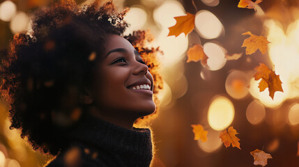 smiling black woman looking up wearing coat in autumn park surrounded by falling leaves golden sunlight bokeh seasons changing fall outdoors natural curly hair smile