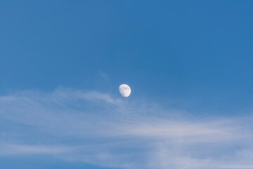 A serene moon captured in a blue daytime sky with soft clouds, conveying calm and peace.