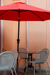 Fototapeta premium A red cloth sun umbrella among round patio tables and white metal bistro chairs. The building exterior wall is orange colored wood. The outdoor street cafe is empty. The sunshade is open in the lounge