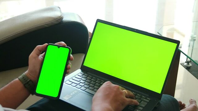 Man using smartphone and laptop with green screens, ideal for custom content or editing.