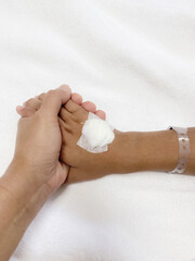 A mother holds the hand of her 9-year-old son who is bedridden. There is agauze on the wound from a...