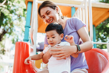 Asian boy and mother playing slider at playground. Together mother and her young son sharing a playful moment on a slide at the park. Asian boy and mother playing slider at playground.