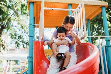 Asian mother and her young son sharing a playful moment on a slide at the park. Together Asian boy...