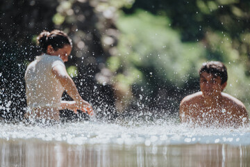 Fototapeta premium Two young adults enjoy a playful moment in water, splashing and laughing under the warm sun in nature.