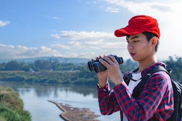 Asian boy wearing a red cap and plaid shirt, looking through binoculars while standing on a bridge over a river. He’s enjoying the natural surroundings and observing wildlife with curiosity.