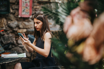 Focused woman using smart phone for work in outdoor cafe with documents and laptop. Outdoor work and remote business concept.