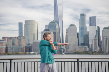 Senior man in sportswear stretching his arms outside in NYC background. Man doing stretching exercise, preparing for workout near Manhattan. Sporty athletic fit man 40s wear sports clothes.