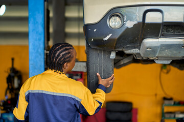 African american female in technicial uniform checking customer car tire whil car on machine power lift in car garage.