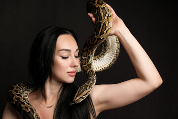 Portrait of a beautiful woman with dark hair on a dark background with a python near her face. Copy...