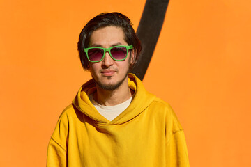 Stylish Asian guy in green glasses stands against a yellow wall,