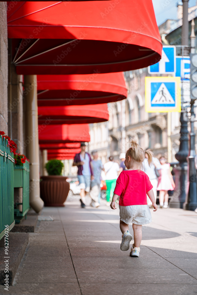 Wall mural A little girl walks along a city street with red canopies of a building in the center of St. Petersburg.