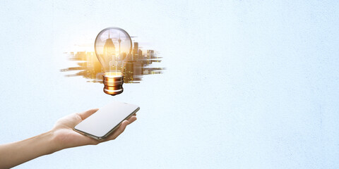 Hand holding smartphone with a lightbulb and cityscape emerging from the screen on a light blue background.