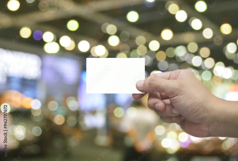 Wall mural Man hand holding blank white business name card in fingers over blur light and shadow of shopping mall