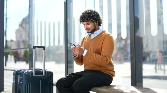 Traveling man sitting on a bench using smartphone at a bus stop. Traveler with suitcase enjoying technology and communication looking at phone screen young man is waiting for transport