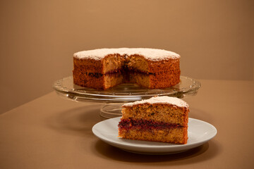 A slice of homemade buckwheat cake (Buchweizentorte, Schwarzplententorte) with raspberry jam filling on a white plate, next to the whole cake on a glass stand
