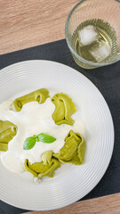 Plate of green tortellini in creamy Alfredo sauce with a sprig of basil alongside a glass of white wine, ideal for Italian cuisine photography