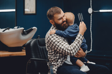 Barber shop, little child is afraid to get haircut, father supports and calms, hugs son in...