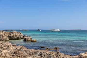 The rocky coastline of Ibiza with yachts anchored in the clear turquoise waters, reflecting a...