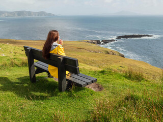 A woman sits on a bench overlooking the ocean. The scene is peaceful and serene, with the woman enjoying the view. Achill island, Ireland. Popular travel and tourism area. Irish nature landscape