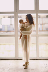 Happy mother and her baby girl in same airy white dresses posing  near window in the studio. Mother holding her little daughter. Copy space for text