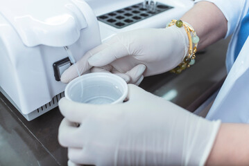 A person wearing gloves holds a cup of distilled water under a semi-auto chemistry analyzer. Aspirating water with a hose. At a medical laboratory or hospital.