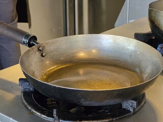 A large metal frying pan with oil heating on a stovetop, preparing for cooking in a kitchen setting. The pan's texture and heat give it a rustic, well-used appearance.

