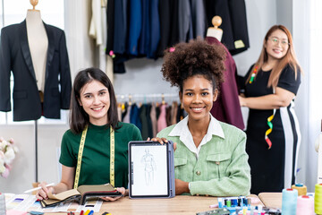 Young two fashion designers use digital tablet meeting with graphic designer discuss show sample dress design new collection on screen on desk while working in sewing workshop