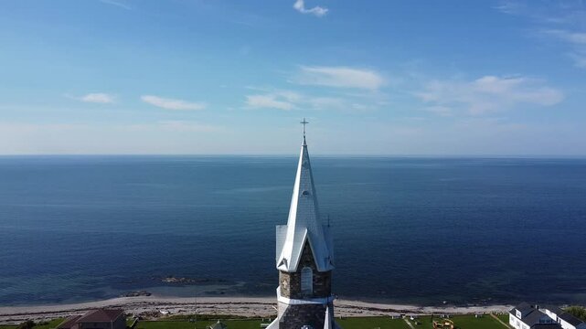 Approche de drone vers un clocher d'&eacute;glise & sa croix de fer forg&eacute; au sommet de sa fl&ecirc;che avec en arri&egrave;re-plan un fleuve d'eau calme se fondant &agrave; l'horizon avec le bleu du ciel et ses nuages. 