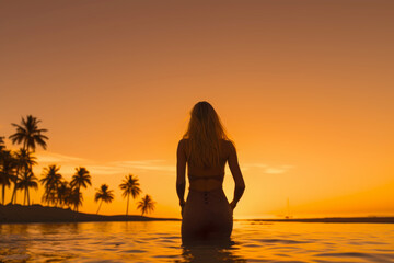 Fototapeta premium The young blonde woman bathes on the beach in the warm sea with palm trees at sunset