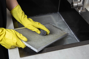 A girl's hands in yellow gloves with a sponge wash the kitchen hood filter with detergent to remove...