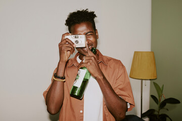 Young black man using silver camera to take a shot for memory while holding bottle of beer