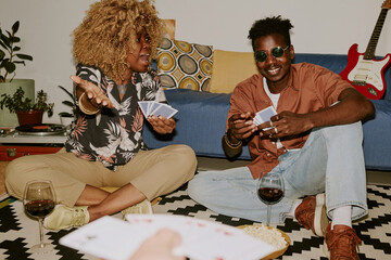Three best friends playing cards on floor while having snacks and drinking wine