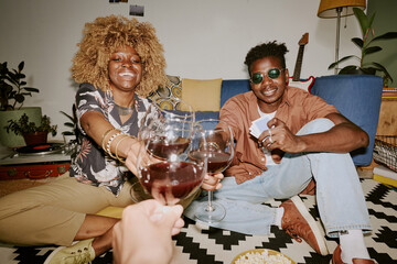 Biracial company of friends drinking red wine and giving toast together while sitting on black...