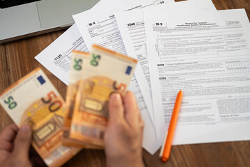 Man freelancer counts euro banknotes preparing for filling various tax forms to report annual income avoiding penalties from government at workplace with laptop