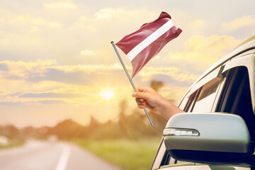 Waving the Latvia flag against the sunrise or sunset from a car driving along a country road. Holding the Latvia flag, traveling by car, on a weekend trip.