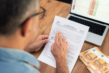 Small business owner examines W-9 tax form to provide Taxpayer Identification Number for reporting income paid by clients at table alongside laptop and euro bills