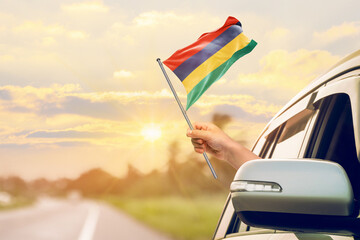 Waving the Mauritius flag against the sunrise or sunset from a car driving along a country road. Holding the Mauritius flag, traveling by car, on a weekend trip.